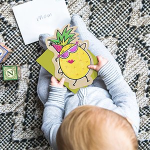 Baby holding a blank note card with a cartoon pineapple wearing sunglasses and a party hat