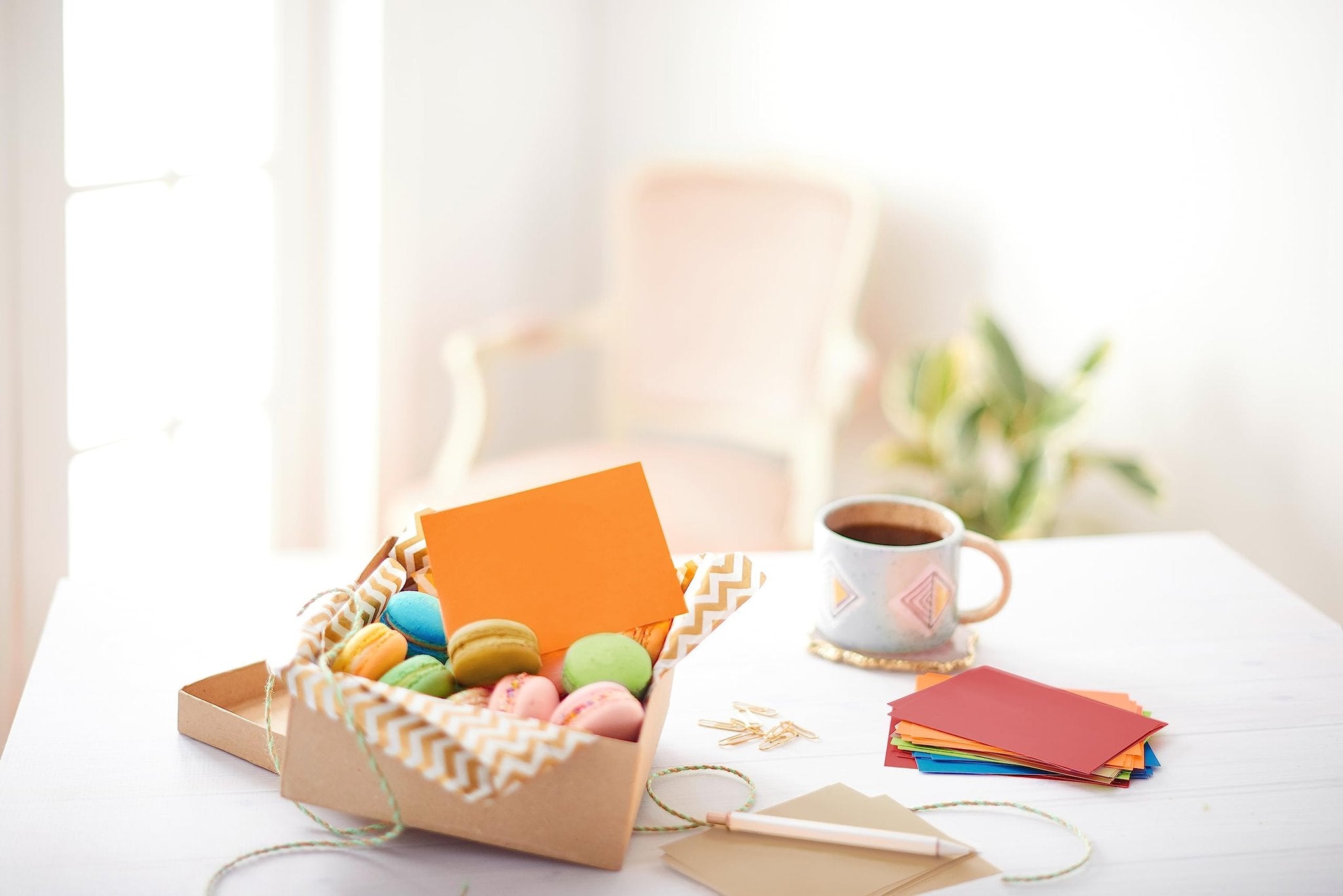 Box of colorful macarons and blank note cards with envelopes, coffee mug, and stationery on a white table.