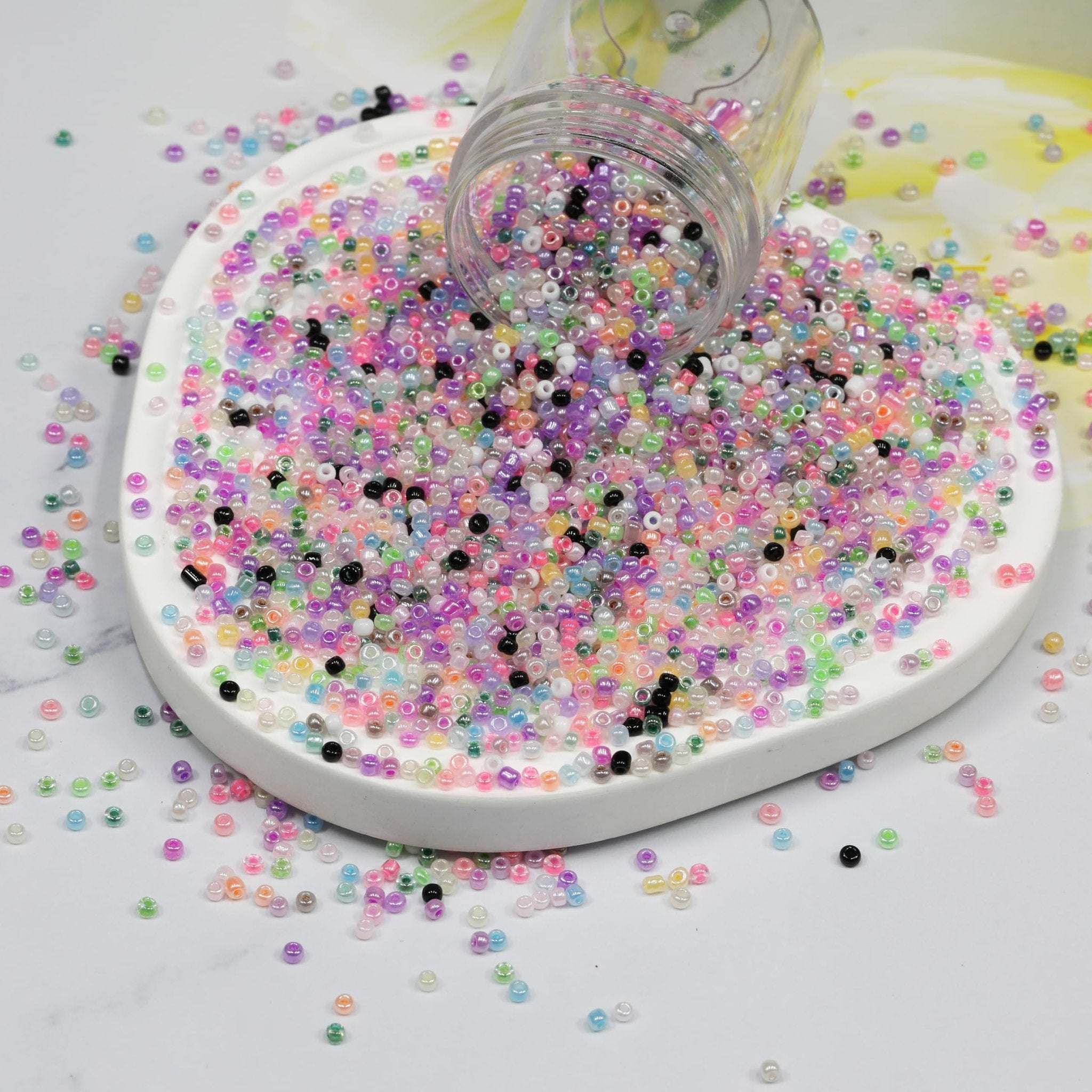 Assorted colorful glass seed beads spilled from a clear container onto a white dish and a white surface.