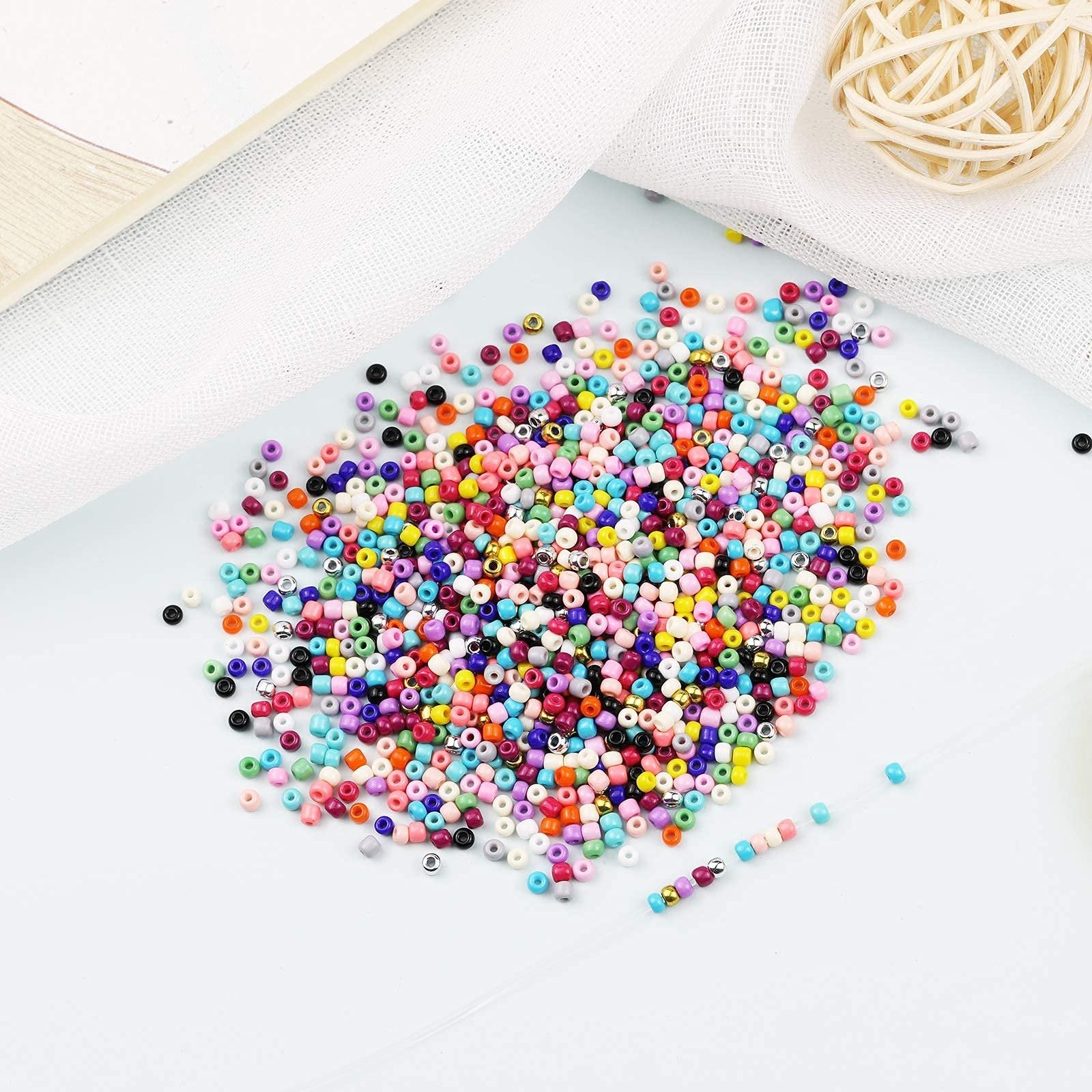 Pile of assorted colorful glass seed beads with gold accents, arranged on a white surface with sheer fabric and a woven sphere.