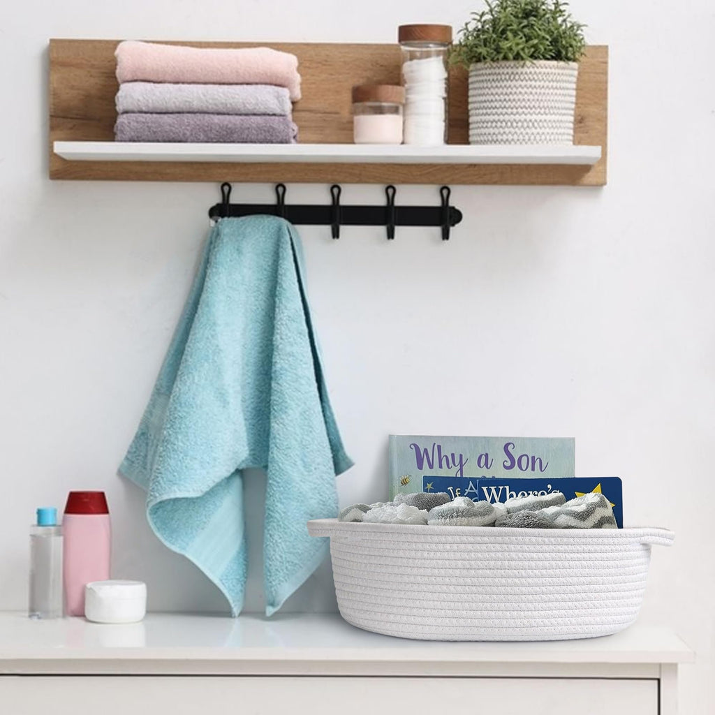 White woven rope basket with handles filled with folded towels on a dresser, next to books and toiletries.