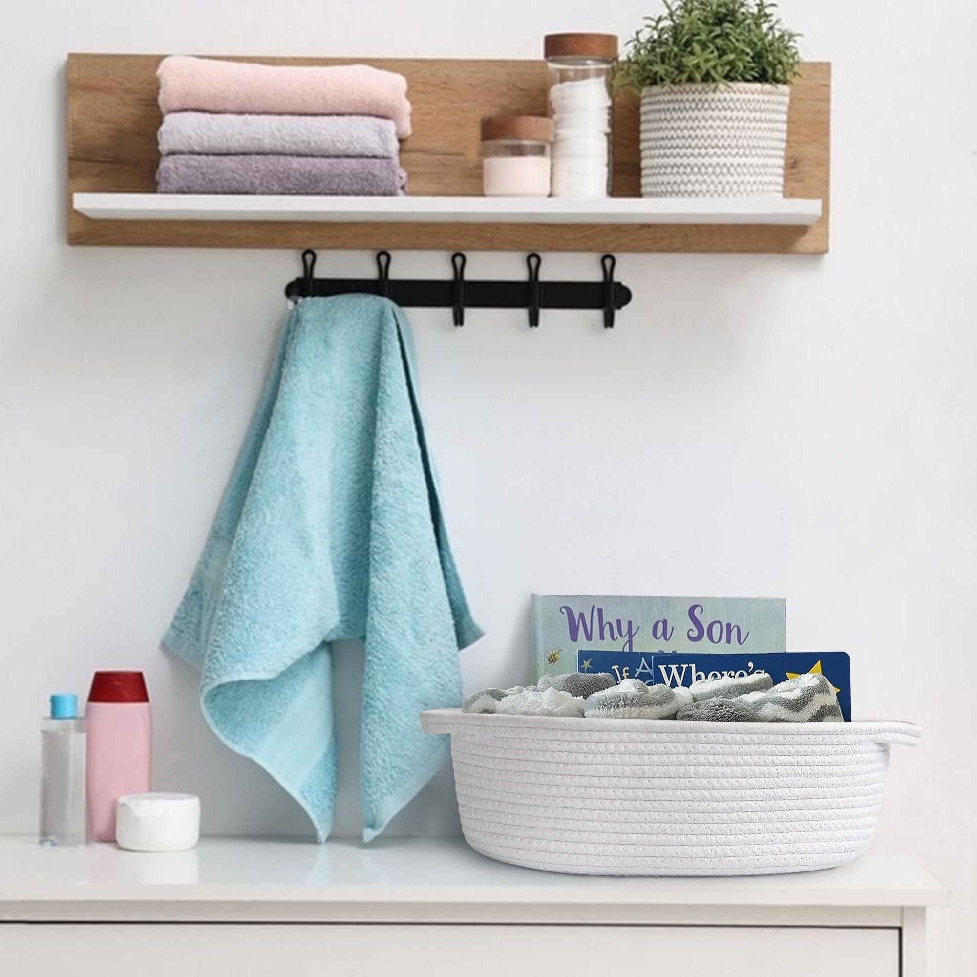 White woven rope basket with handles filled with folded towels on a dresser, next to books and toiletries.