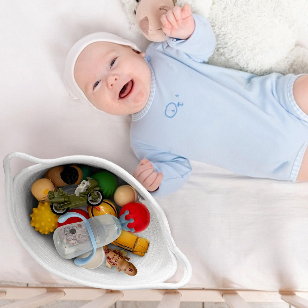 Baby lying on white bedding next to a woven rope basket filled with toys and a baby bottle