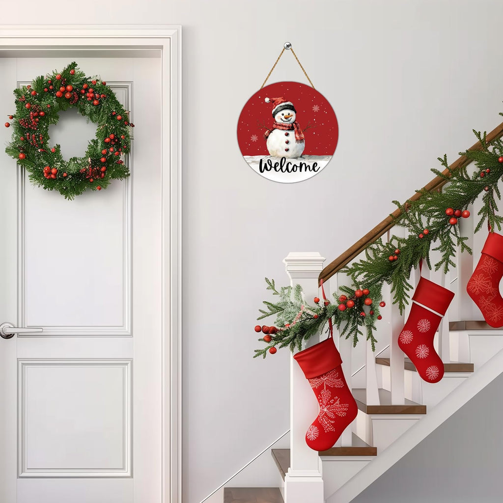 Round wooden snowman welcome door hanger with red background, hanging on a wall next to a Christmas wreath and decorated staircase.
