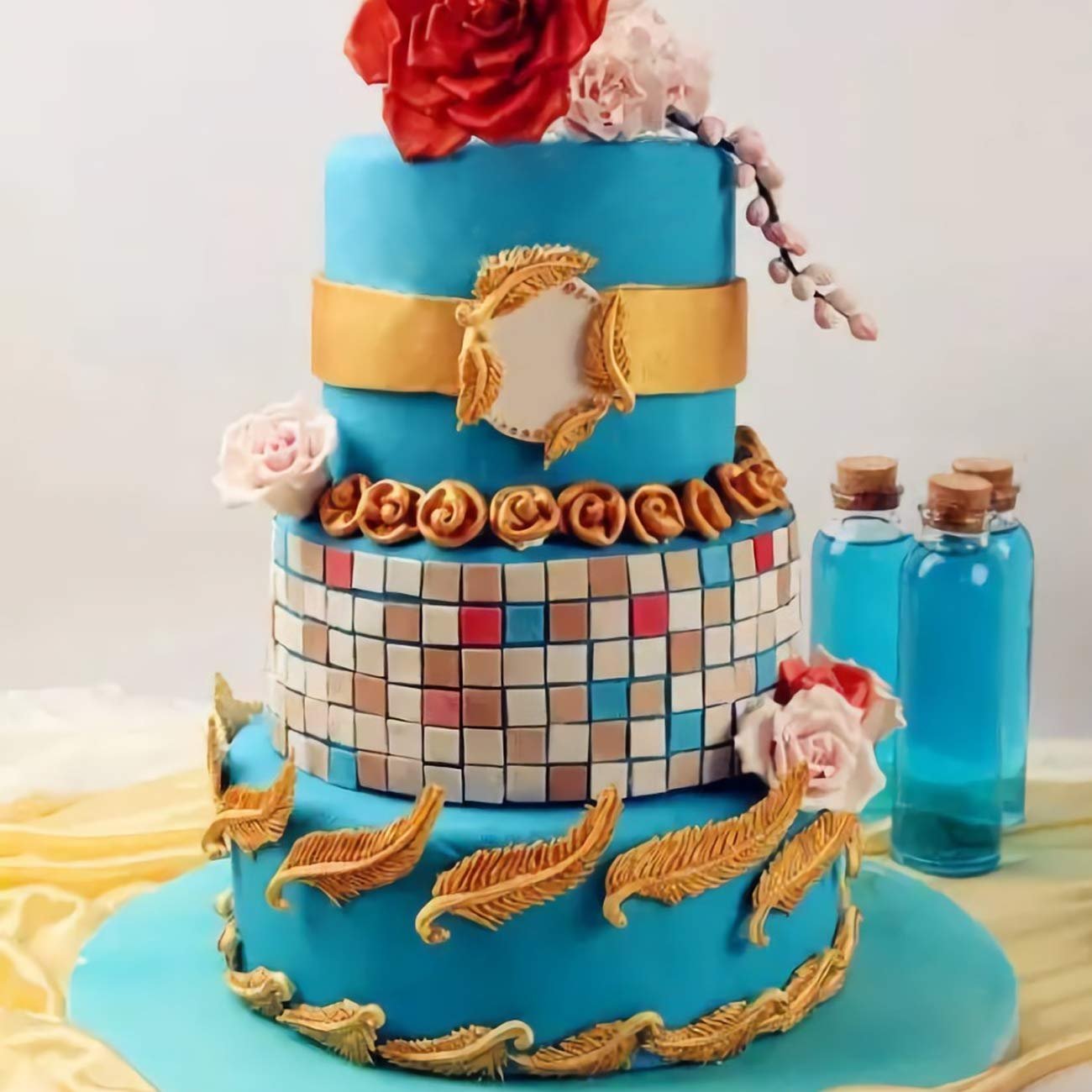 Three-tier blue cake decorated with gold feathers, mosaic pattern, and red and pink flowers, with blue bottles in the background.