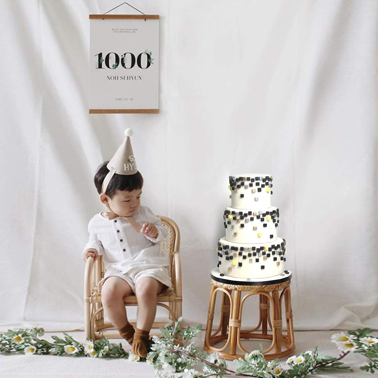 Toddler in party hat sits by a tiered cake decorated with black and gold squares.