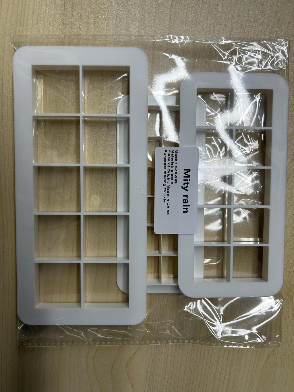 Two white square cookie cutters with grid patterns, packaged in clear plastic on a wooden surface.
