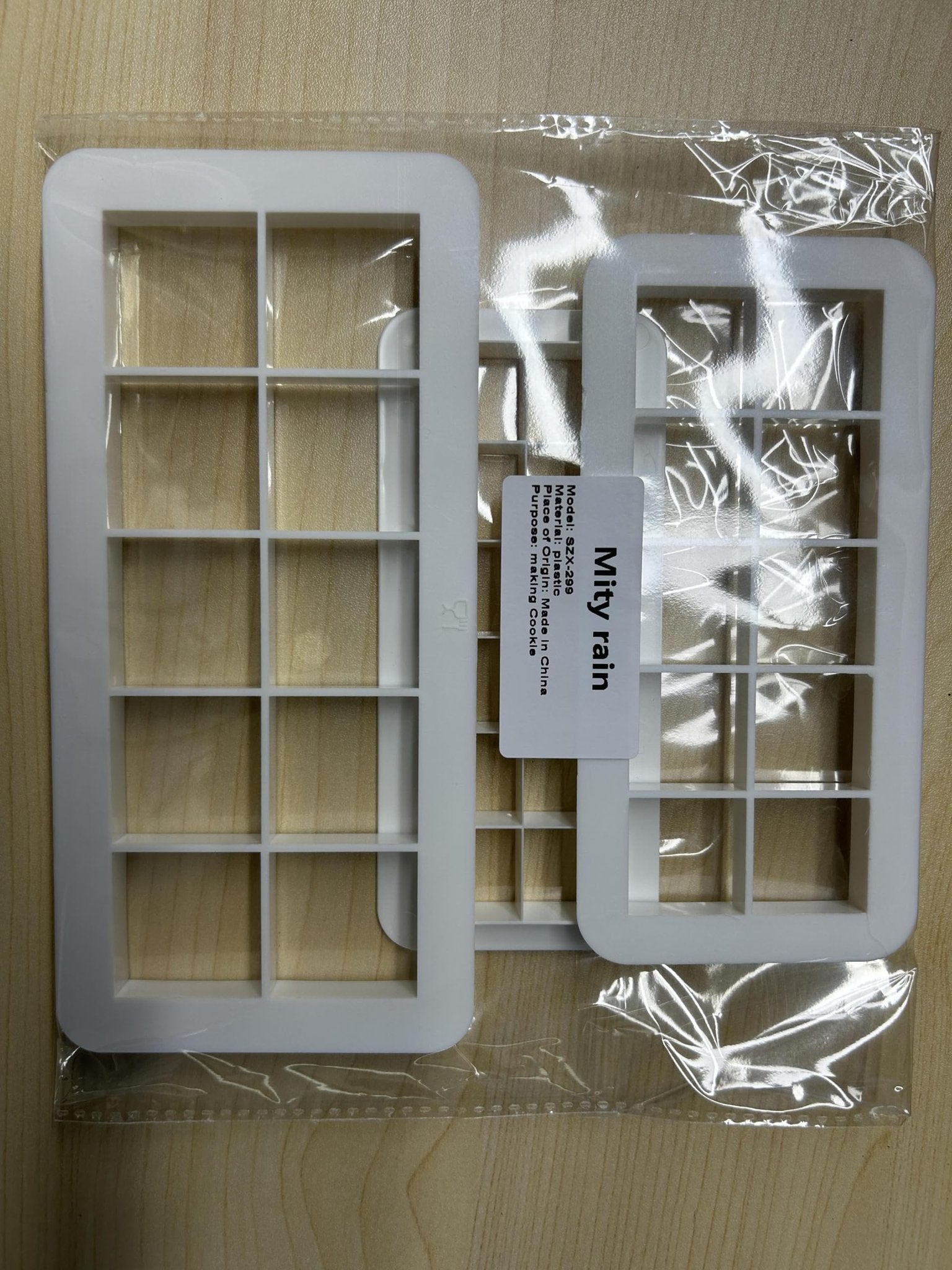 Two white square cookie cutters with grid patterns, packaged in clear plastic on a wooden surface.