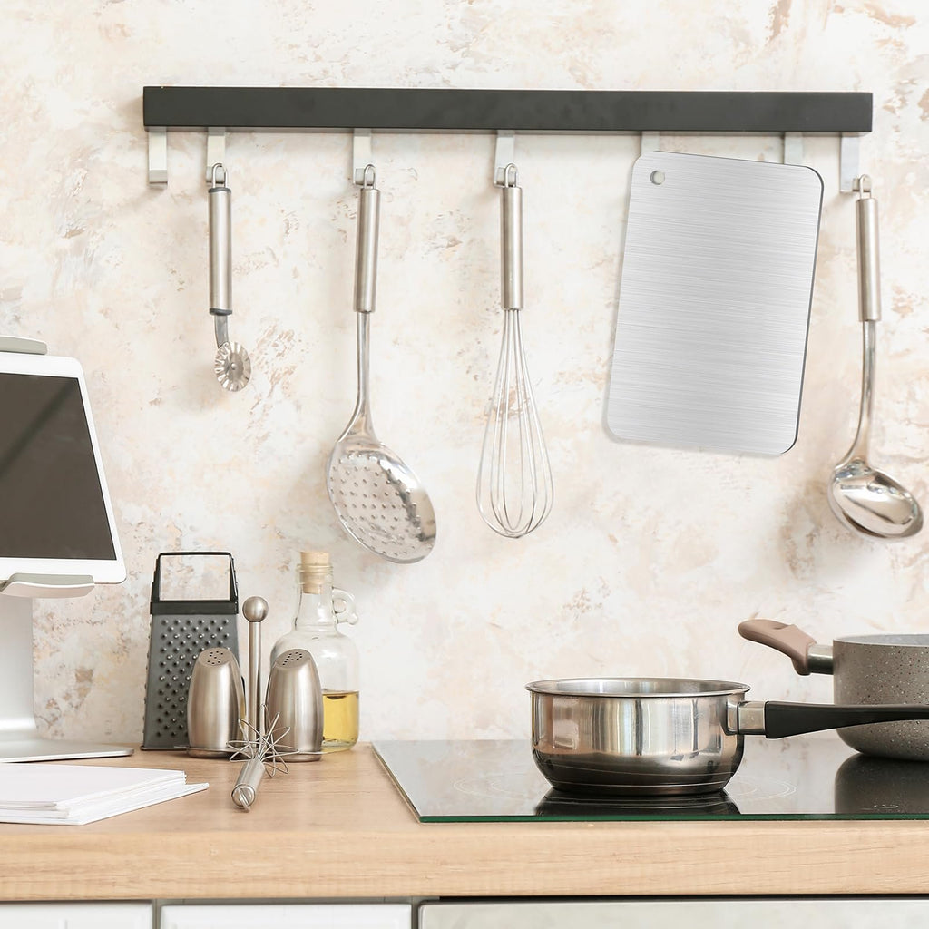 Stainless steel cutting board hanging on a kitchen wall rack with utensils, next to a pot on a stovetop.