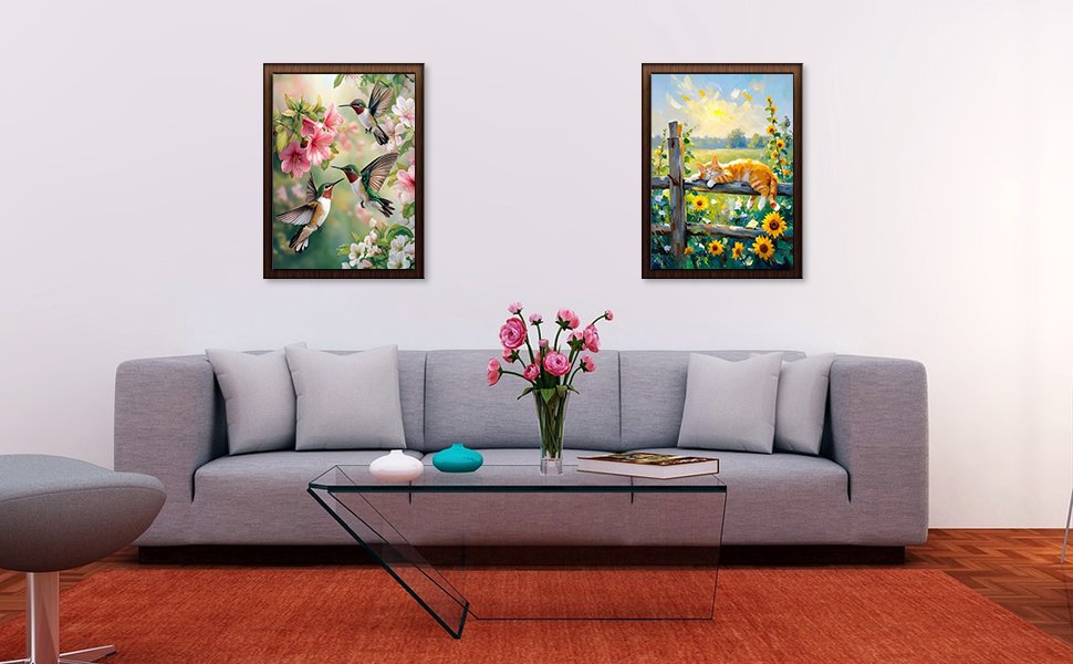 Living room scene with two framed cross stitch art pieces hanging above a gray sofa, featuring hummingbirds and a sleeping cat.
