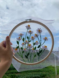 Hand holding an embroidery hoop with a finished daisy pattern against a cloudy sky background.