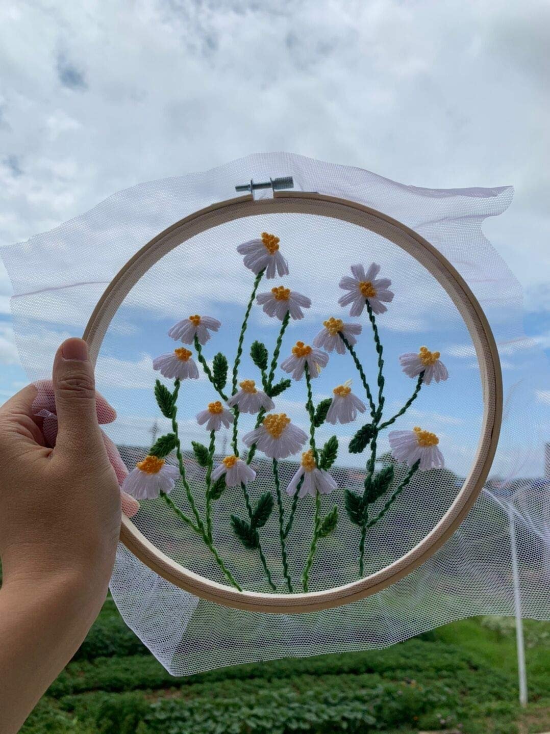 Hand holding an embroidery hoop with a finished daisy pattern against a cloudy sky background.