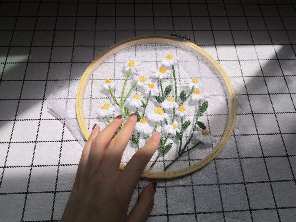 Embroidery hoop with white daisies and green leaves stitched on white fabric, held by a hand.