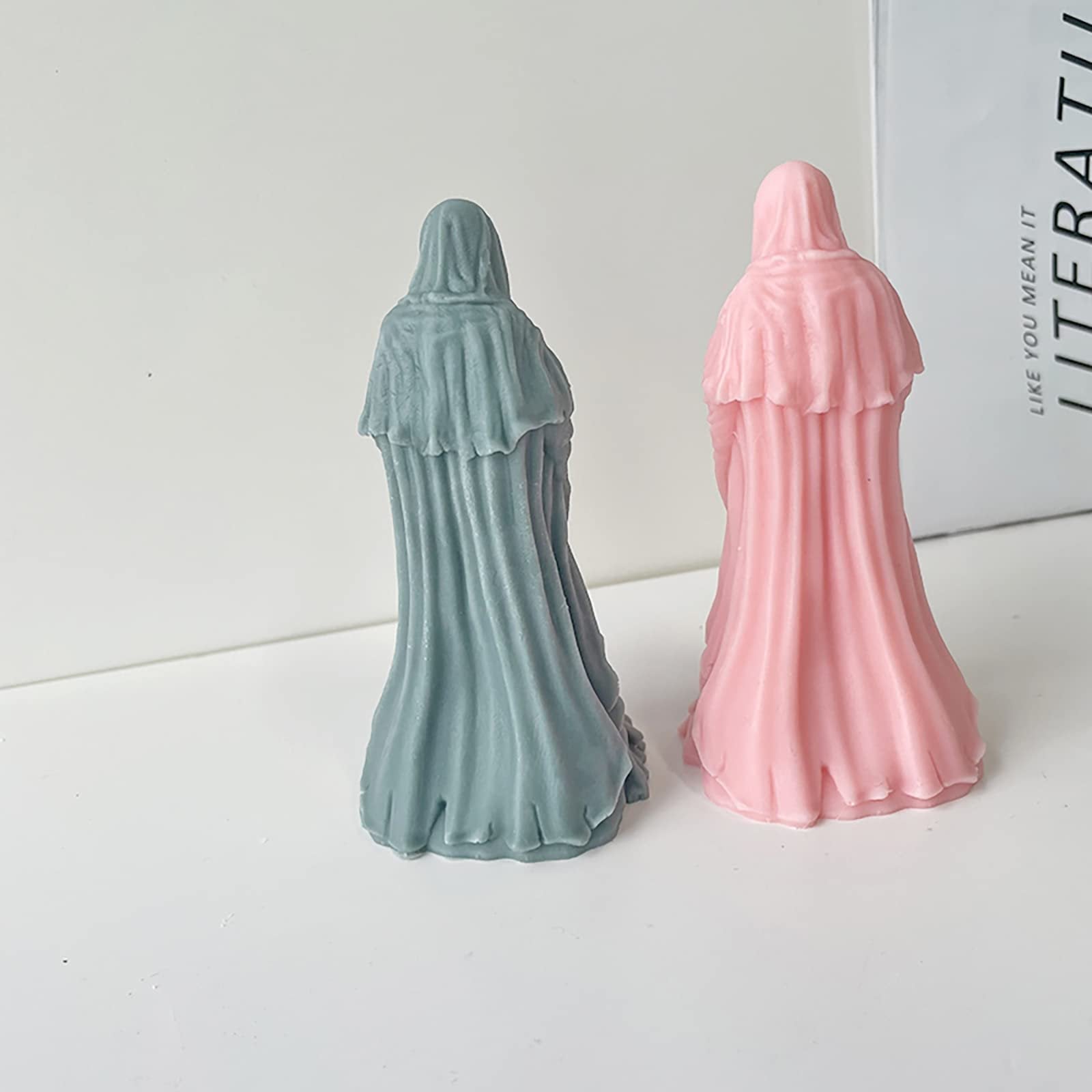 Two hooded figures, one grey and one pink, stand facing away from the camera on a white surface with a book in the background.