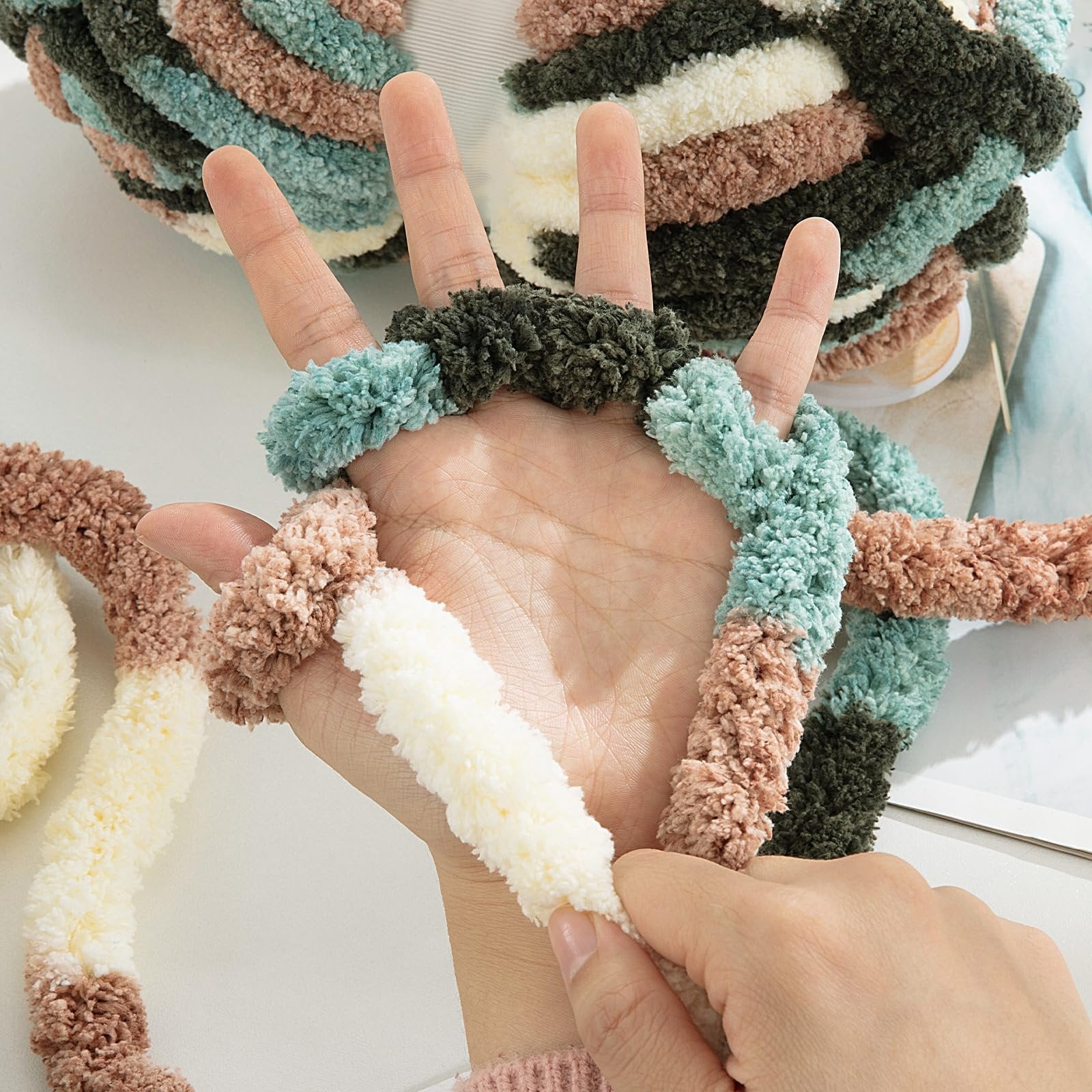 Hand demonstrating chunky chenille yarn in apricot green, brown, cream, and dark green stripes