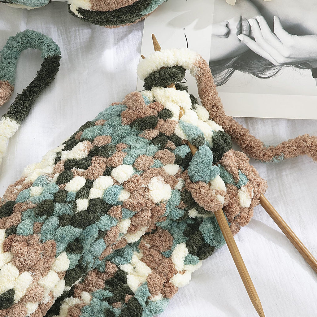 Chunky chenille yarn in apricot green tie-dye pattern being knitted with wooden needles on a white surface.