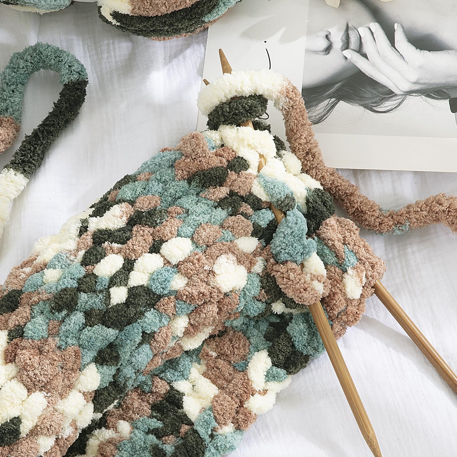 Chunky chenille yarn in apricot green tie-dye pattern being knitted with wooden needles on a white surface.