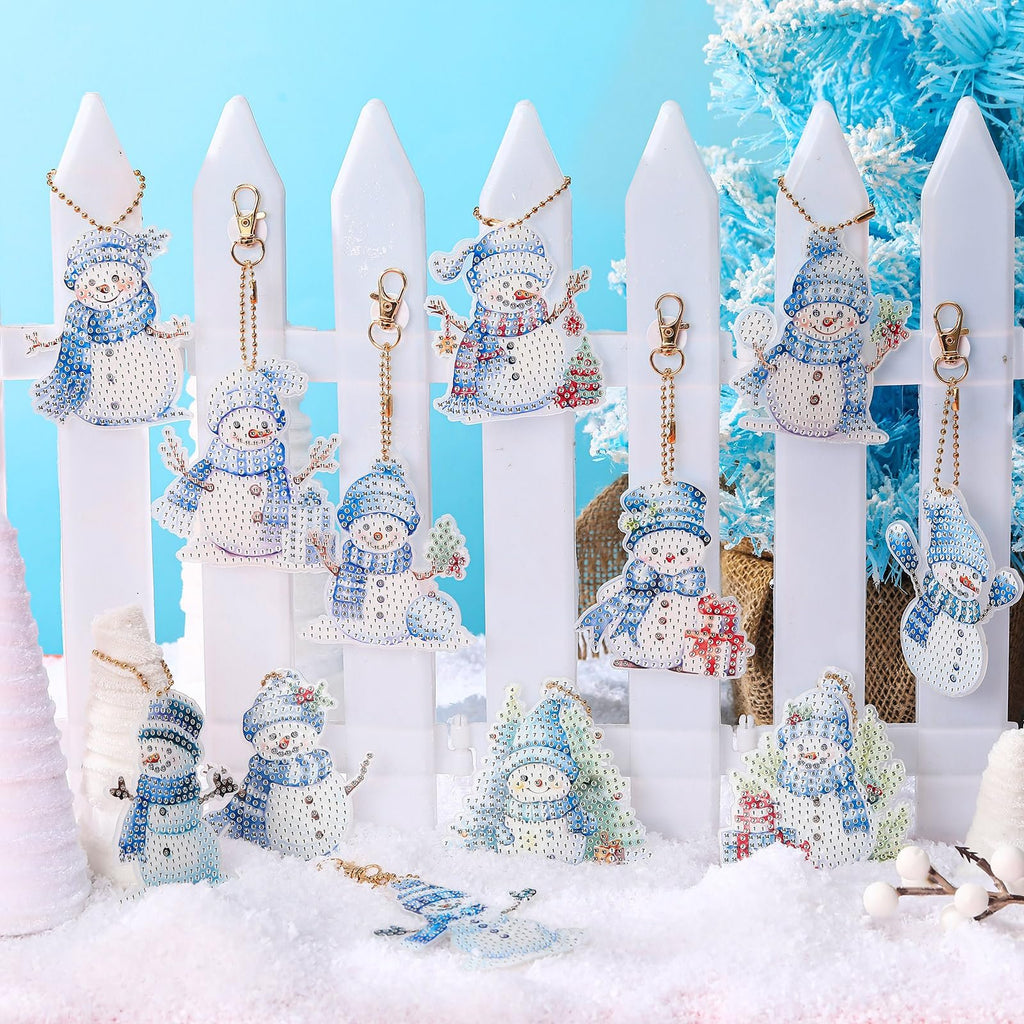 Collection of twelve diamond art snowman keychain ornaments displayed on a white picket fence with faux snow and a blue background.