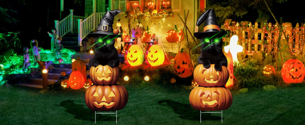 Two black cat Halloween yard signs stacked on pumpkins with witch hats and glowing green eyes, set in a decorated yard at night.