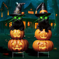 Two black cats wearing witch hats sit atop stacked jack-o'-lanterns in front of a haunted house, with green laser eyes.