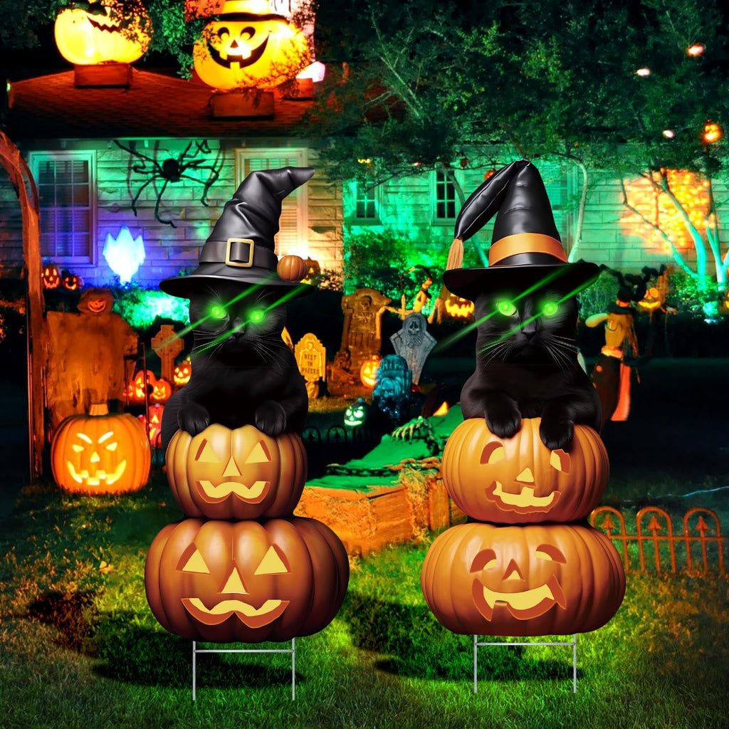 Two black cat Halloween yard signs with witch hats and glowing green eyes, stacked on carved pumpkins, in a decorated yard at night.