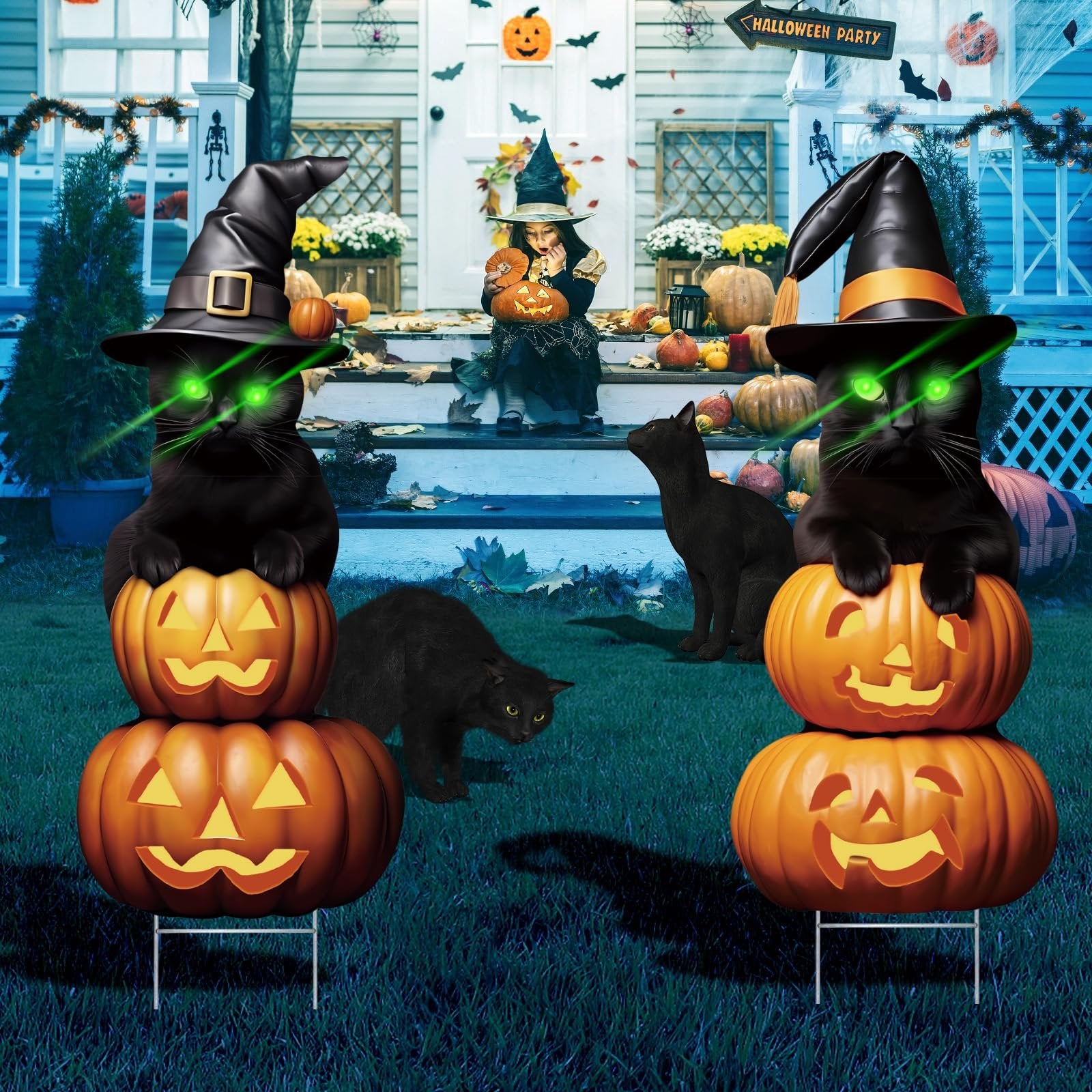 Two black cat Halloween yard signs with glowing green eyes, wearing witch hats, stacked on carved pumpkins with stakes, in front of a decorated porch.