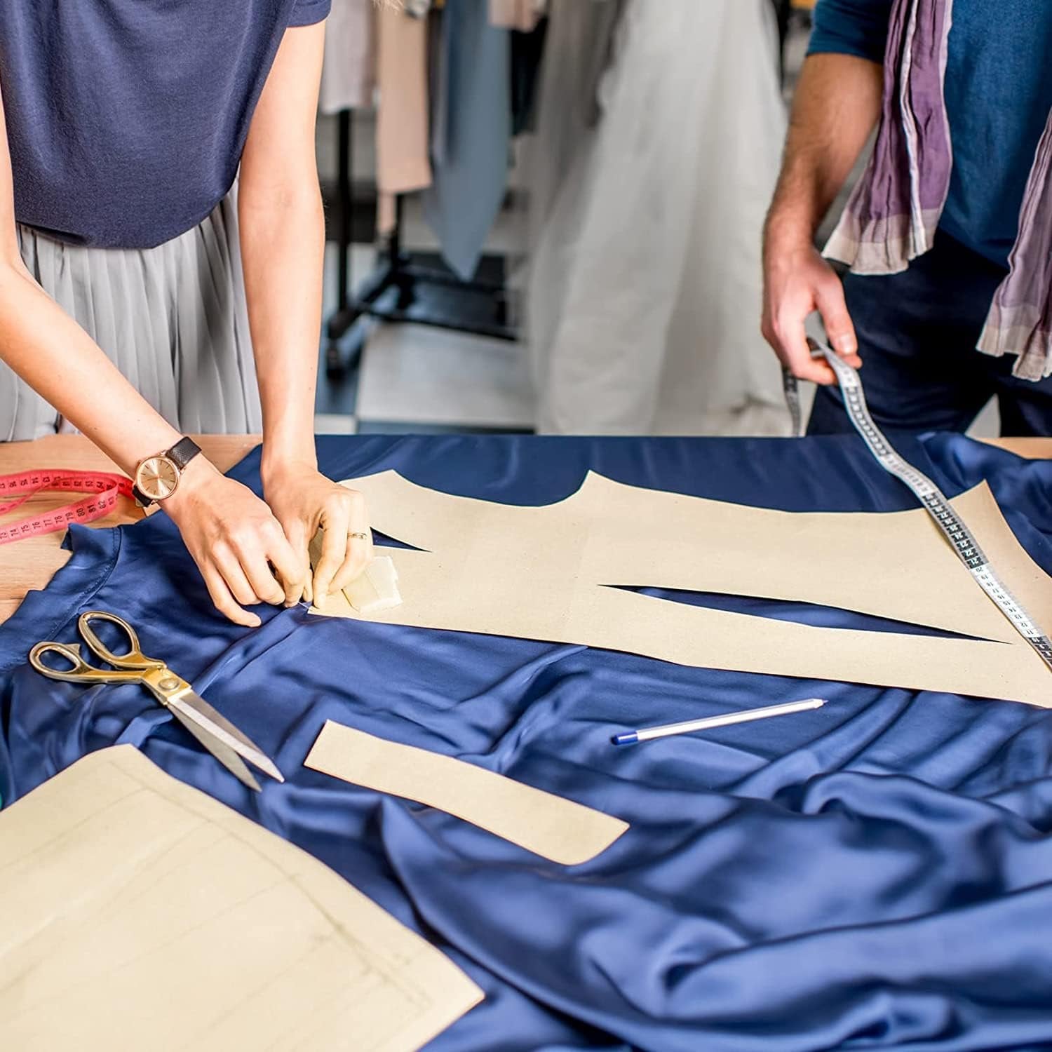 Two people working with fabric and patterns on a table, one pinning a pattern piece to blue satin fabric.