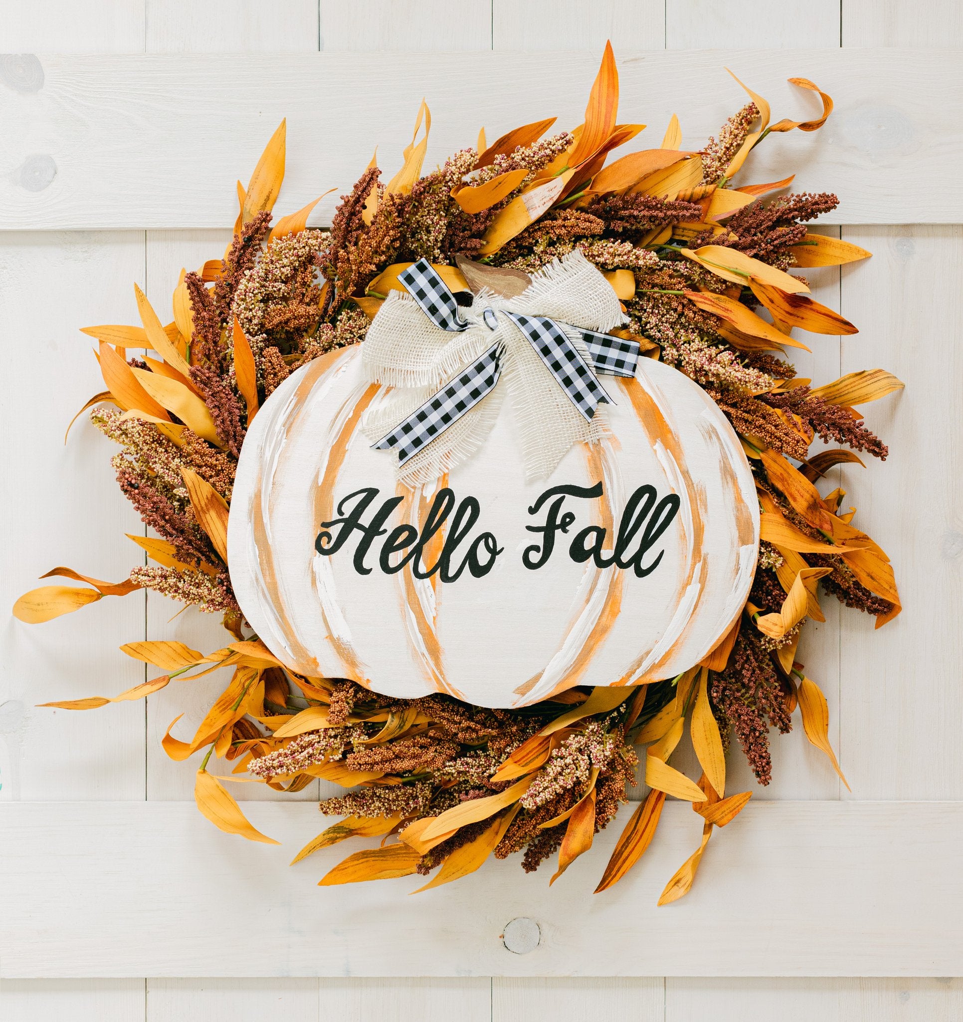 White painted pumpkin cutout with 'Hello Fall' text, decorated with a burlap bow and autumn foliage, against a white wooden background.