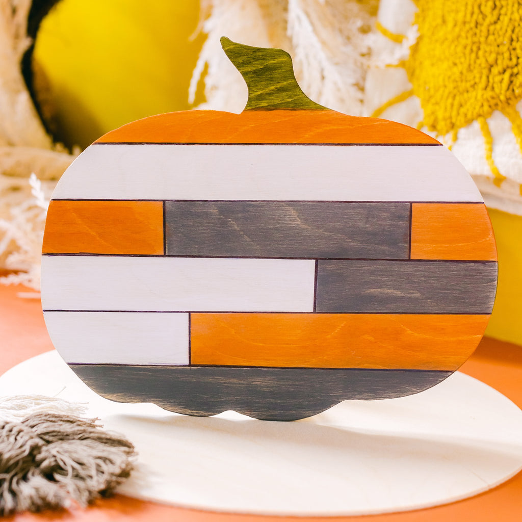 Wooden pumpkin cutout with horizontal stripes of orange, white, and gray, set against a blurred background of yellow and white.