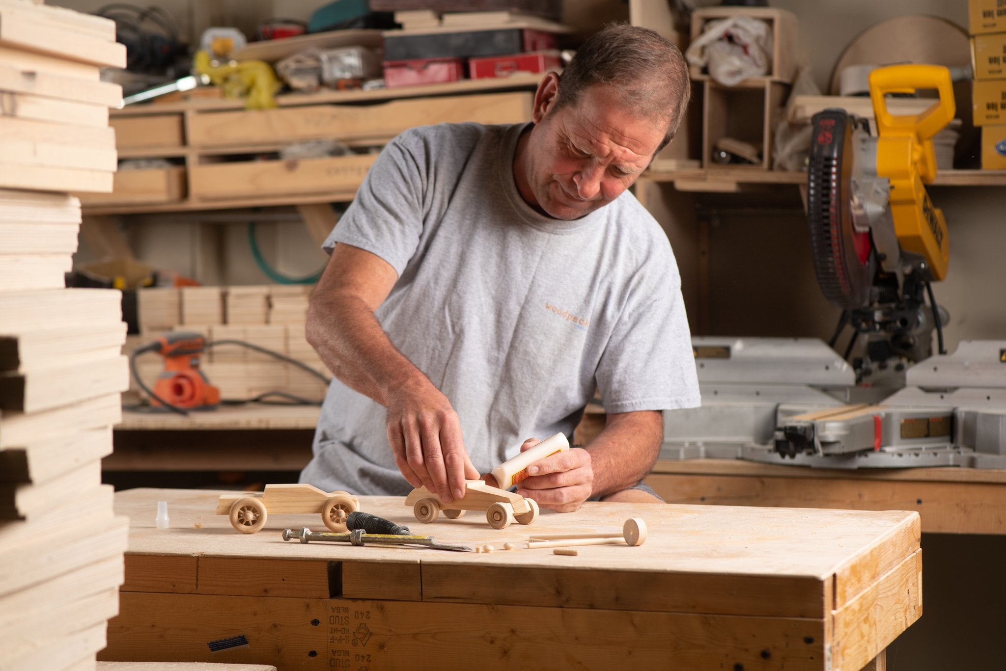 Man assembling wooden toy cars with glue in a workshop setting, stacks of wood in foreground