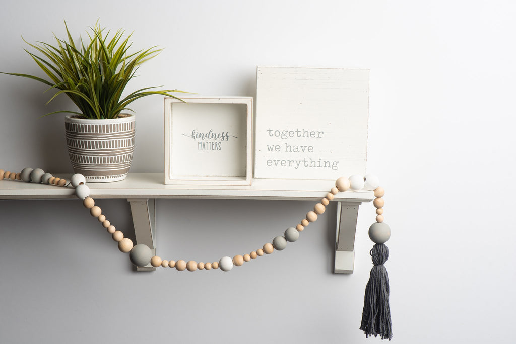 Shelf decorated with a potted plant, two decorative signs, and a beaded garland with a tassel.