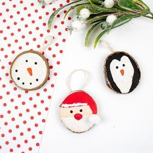 Three unfinished wood slice ornaments: a snowman, a penguin, and Santa Claus, arranged on a white surface with red polka dots and greenery.