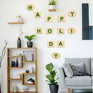 Wooden letters spelling 'Happy Holi Day' on a white wall above a wooden shelf and grey sofa.