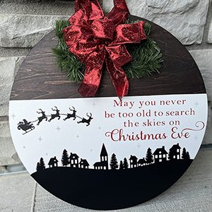 Round wooden Christmas decoration with a village silhouette, Santa's sleigh, and text, topped with a red bow and greenery.