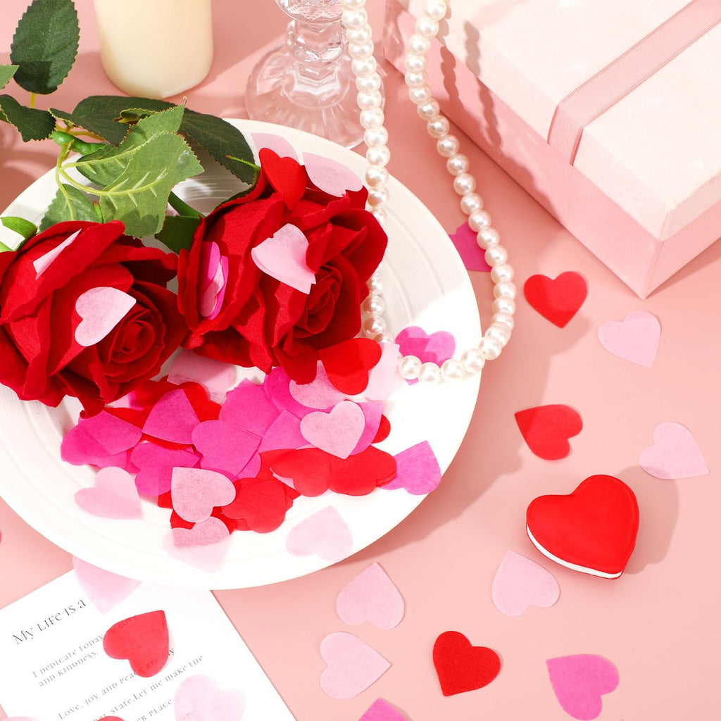 Valentine's Day heart confetti in red, rose, and pink scattered around two red roses on a white plate, with a pearl necklace and gift box.