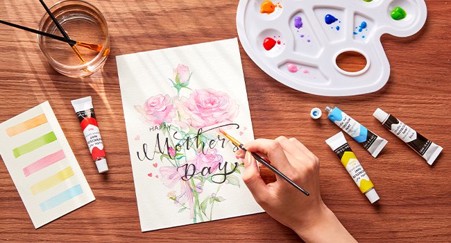 Hand painting a "Happy Mother's Day" card with pink roses using watercolor paints and brushes.