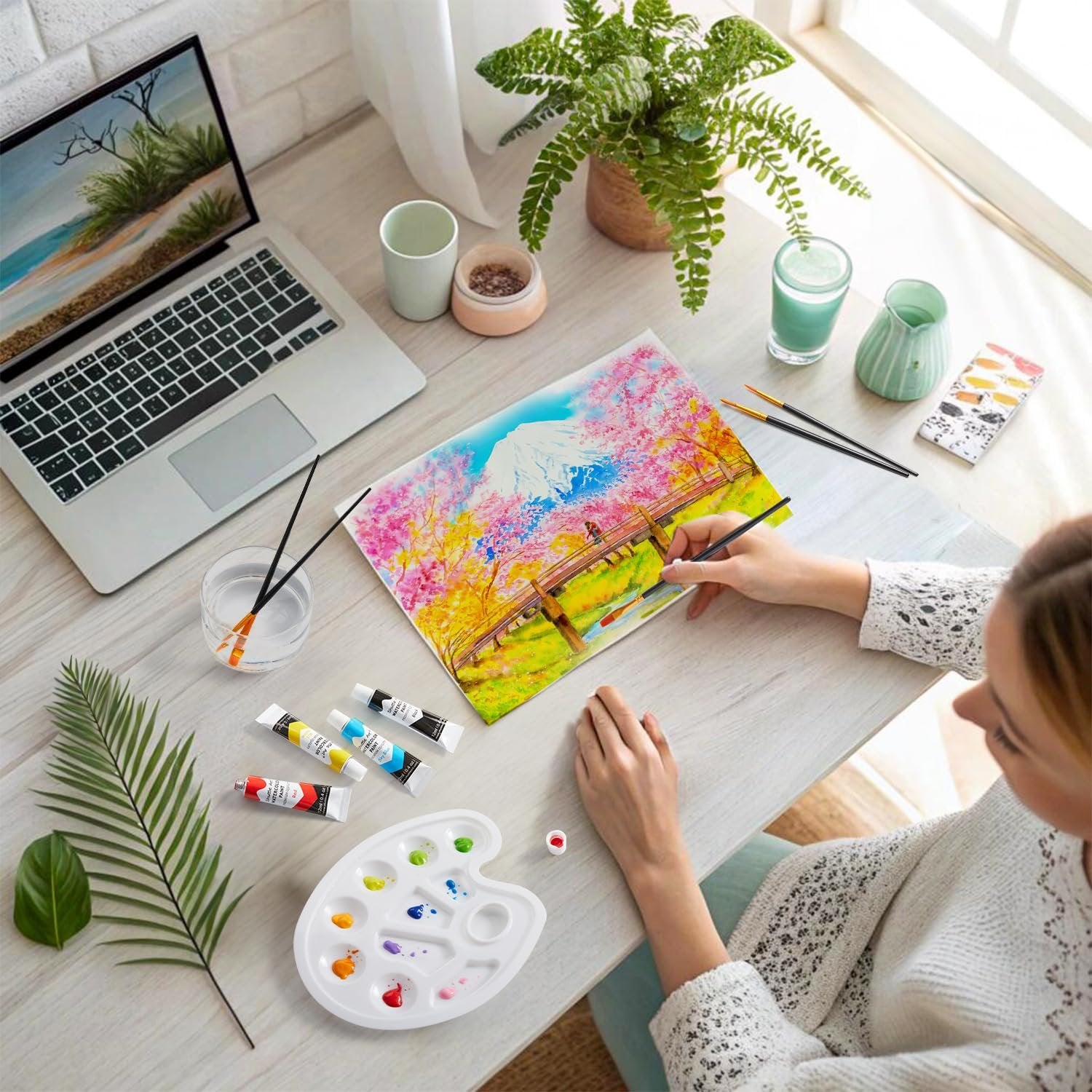 Person painting a landscape with watercolor paints and brushes at a desk with a laptop