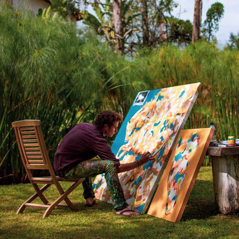 Artist painting on a large canvas outdoors with a wooden chair and table nearby