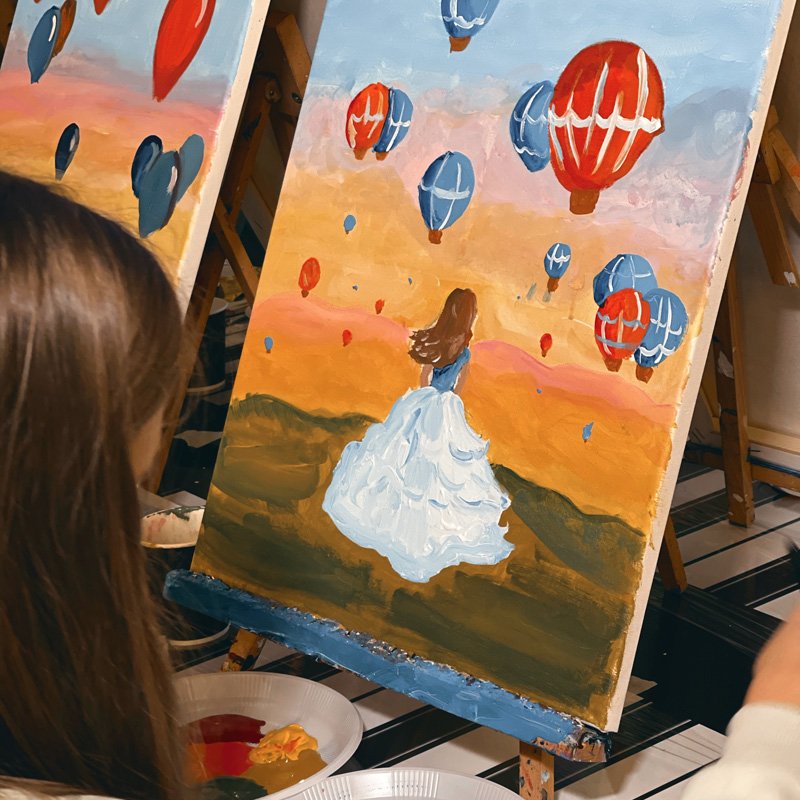 Woman painting a hot air balloon landscape on canvas at an art class