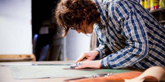 Person wearing glasses and a blue plaid shirt cuts material with a craft knife on a cutting mat.