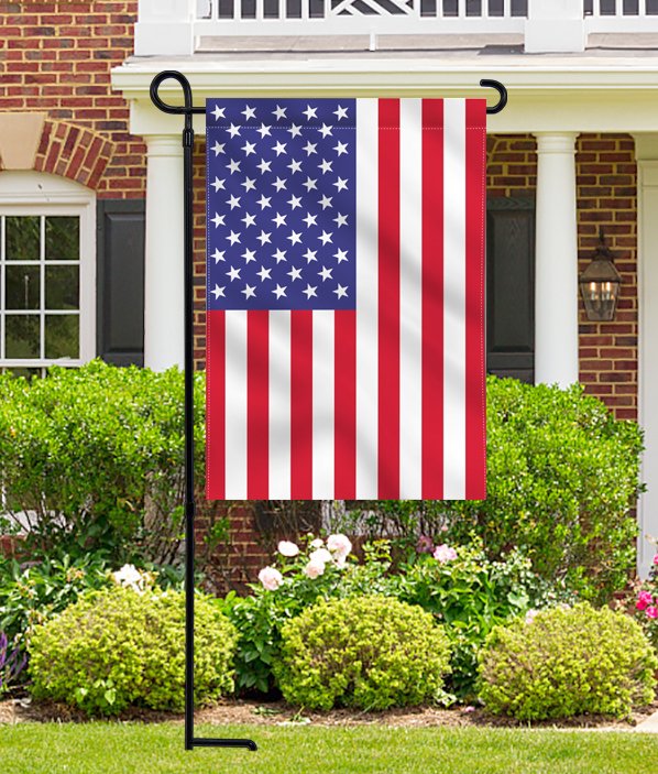 Black metal garden flag stand holding an American flag in front of a brick house with landscaping.