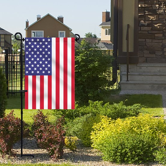 Black metal garden flag stand with an American flag displayed in a yard with houses in the background.