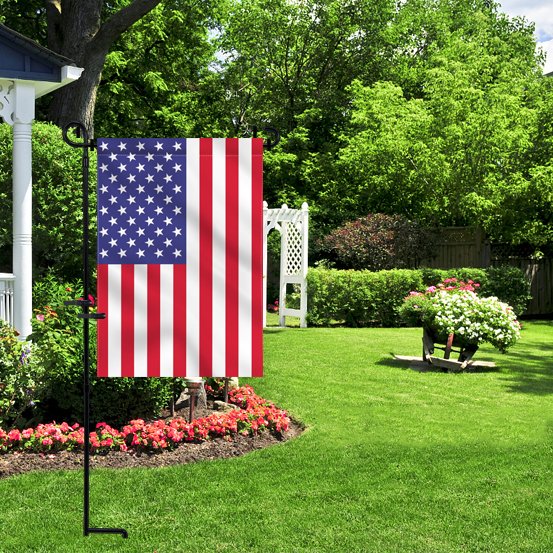 American flag on a black metal stand in a lush green garden with flowers and trees.