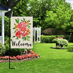 Black metal garden flag stand holding a welcome flag with a floral design, set in a lush green yard with a white arbor and flower beds.