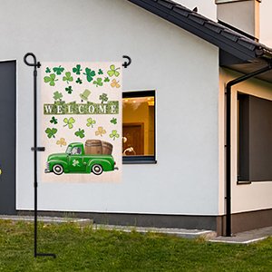 Black metal garden flag stand holding a St. Patrick's Day flag with a green truck and shamrocks, placed in a yard next to a house.
