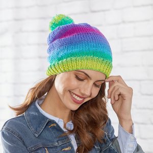 Woman wearing a colorful knitted beanie with a pompom, against a white brick wall background.