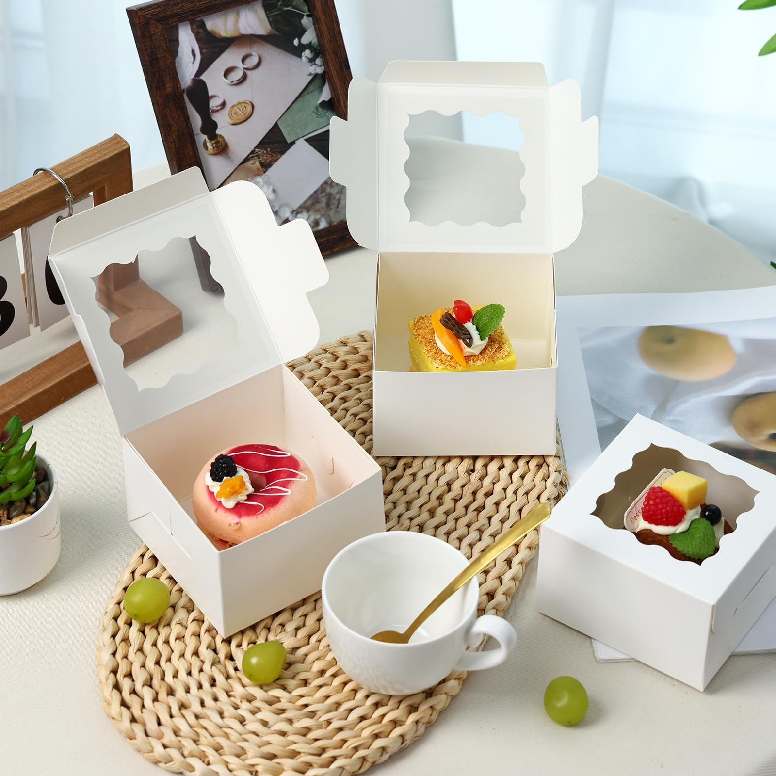 Three white bakery boxes with clear windows, one containing a donut and two containing small cakes, arranged on a table with grapes and a teacup.