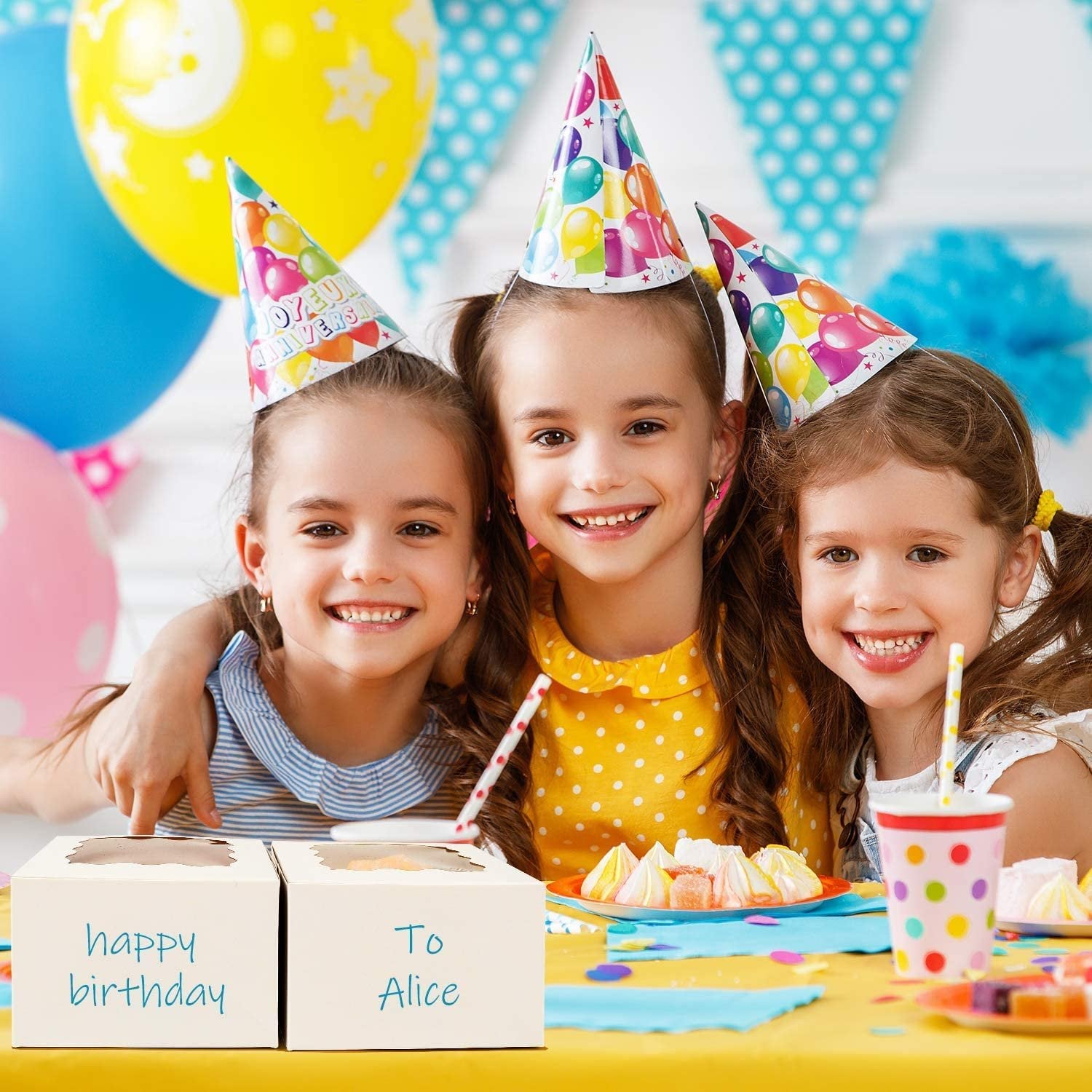 Three smiling girls in party hats at a birthday table with two white cookie boxes, one labeled 'happy birthday' and the other 'To Alice'.