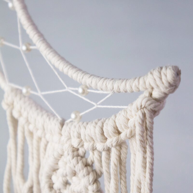 Close-up of a white macrame dream catcher with pearl beads and tassels against a light blue background.