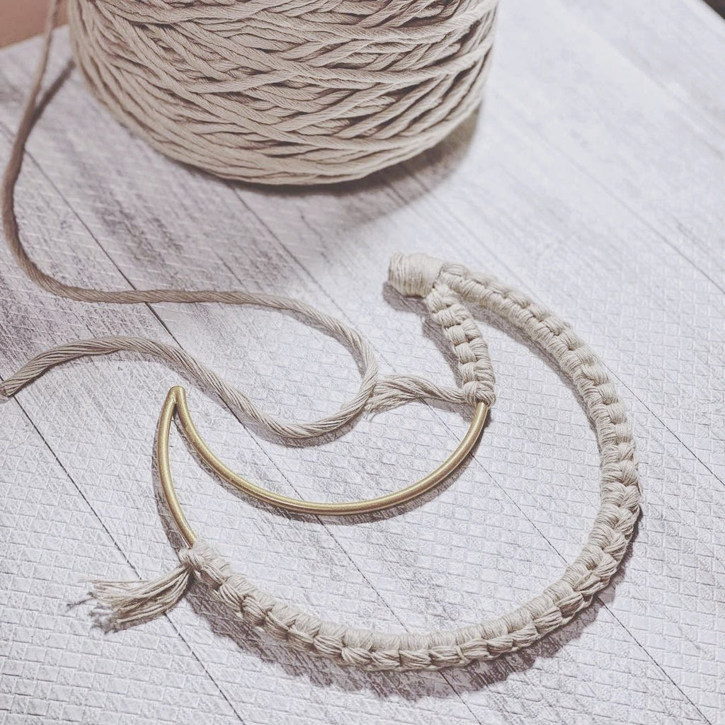 Macrame crescent moon dream catcher in progress with a large spool of cotton cord on a light wood-grain surface.